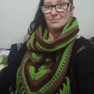 May include: A green and brown crocheted triangular scarf with fringe. The scarf is worn by a person with dark hair and glasses.