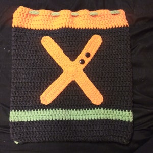 May include: A crocheted bag with a black background, orange 'X' and green trim. The bag has two black buttons on the 'X'.
