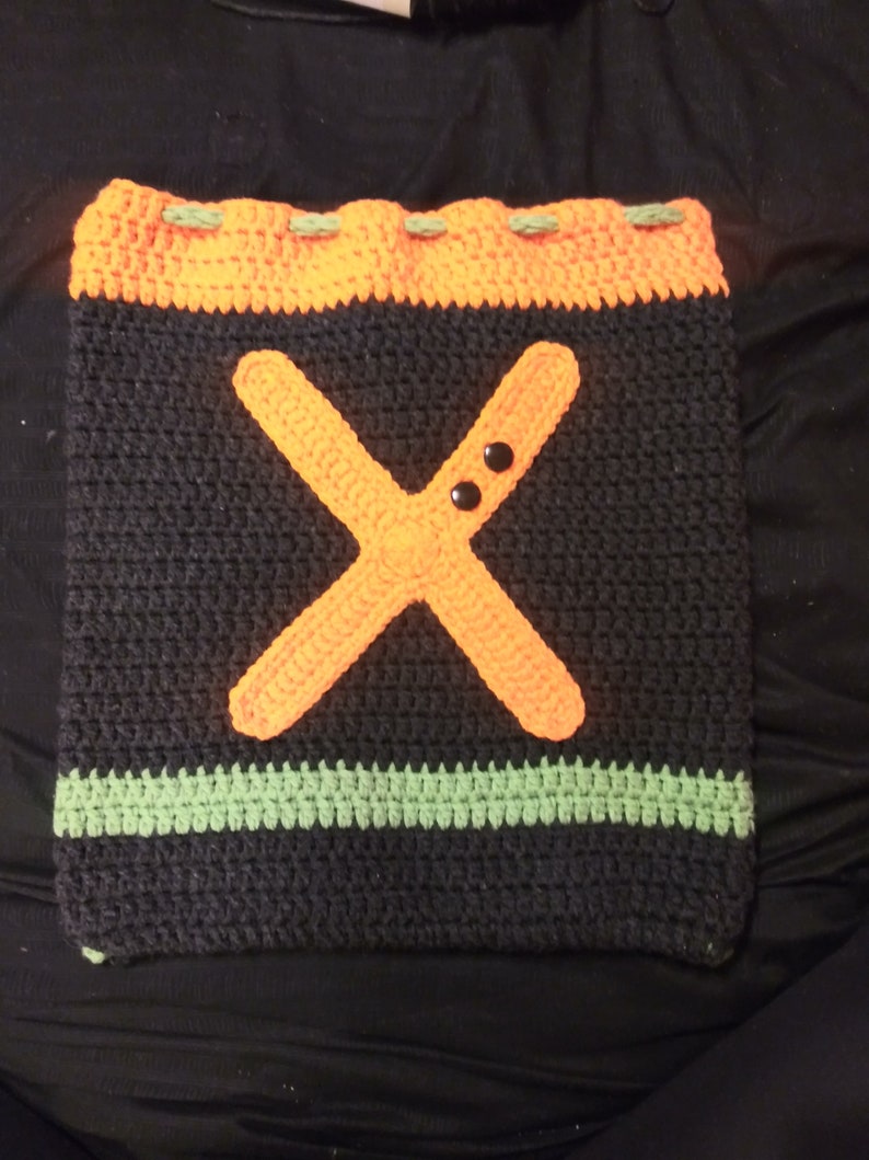 May include: A crocheted bag with a black background, orange 'X' and green trim. The bag has two black buttons on the 'X'.