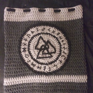 May include: A grey and black crocheted bag with a circular design featuring a black triangle symbol surrounded by runes. The bag has a drawstring closure.