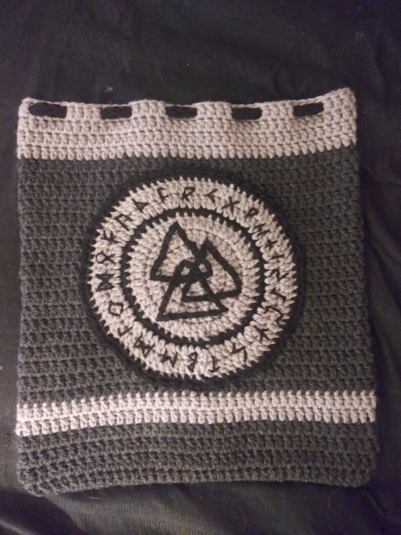 May include: A grey and black crocheted bag with a circular design featuring a black triangle symbol surrounded by runes. The bag has a drawstring closure.