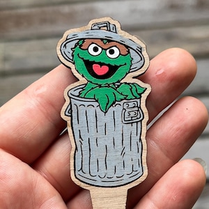 Pode incluir: Estaca de jardim de madeira com Oscar, o Gruñón, da Rua Sésamo. A personagem é retratada no seu caixote do lixo, com um corpo verde, um caixote do lixo cinzento e uma tampa cinzenta. A estaca é feita de madeira clara.