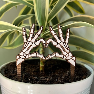 May include: Wooden skeleton hands forming a heart shape, a plant pot with soil and a green plant with white stripes in the background.