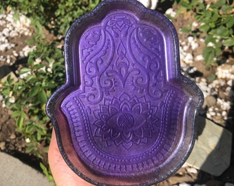 May include: Purple resin hand-shaped dish with an intricate design and a raised eye in the center.