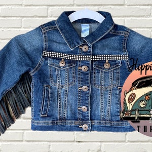 Peut inclure: Une veste en jean bleue avec des franges sur les manches et une rang&eacute;e de strass sur la poitrine. La veste a une fermeture &agrave; boutons et deux poches. L'image comprend &eacute;galement un logo avec un van vintage et le texte "Hippie Misfit Threads".