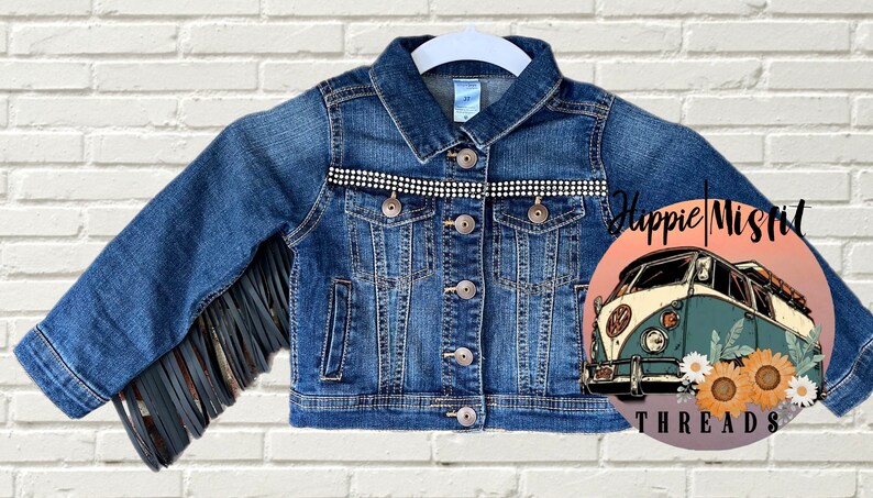 Peut inclure: Une veste en jean bleue avec des franges sur les manches et une rang&eacute;e de strass sur la poitrine. La veste a une fermeture &agrave; boutons et deux poches. L'image comprend &eacute;galement un logo avec un van vintage et le texte "Hippie Misfit Threads".