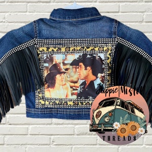 Peut inclure: Une veste en jean bleue avec des franges noires sur les manches et une bordure de strass. Le dos de la veste pr&eacute;sente une photo d'un couple dans un d&eacute;cor western, encadr&eacute;e d'un motif l&eacute;opard et de strass. La veste est d&eacute;cor&eacute;e du texte "Hippie Misfit Threads".