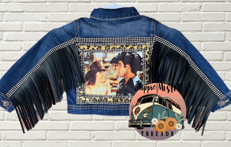 Peut inclure: Une veste en jean bleue avec des franges noires sur les manches et une bordure de strass. Le dos de la veste pr&eacute;sente une photo d'un couple dans un d&eacute;cor western, encadr&eacute;e d'un motif l&eacute;opard et de strass. La veste est d&eacute;cor&eacute;e du texte "Hippie Misfit Threads".