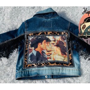 Peut inclure: Une veste en jean bleue avec des franges noires et un patch orn&eacute; de strass repr&eacute;sentant un couple sur une photo vintage. Le patch est entour&eacute; d'un motif l&eacute;opard. La veste porte le texte "Hippie Misfit Threads" au dos.