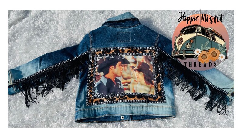 Peut inclure: Une veste en jean bleue avec des franges noires et un patch orn&eacute; de strass repr&eacute;sentant un couple sur une photo vintage. Le patch est entour&eacute; d'un motif l&eacute;opard. La veste porte le texte "Hippie Misfit Threads" au dos.