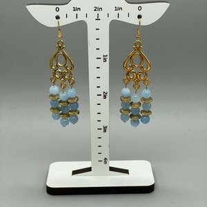 Puede incluir: Pendientes de aro dorados con cuentas de color azul claro y un diseño de filigrana. Los pendientes miden aproximadamente 5 cm de largo.