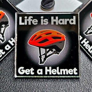 May include: Three black and silver enamel pins with a black and orange bicycle helmet graphic. The pins say "Life is Hard Get a Helmet".