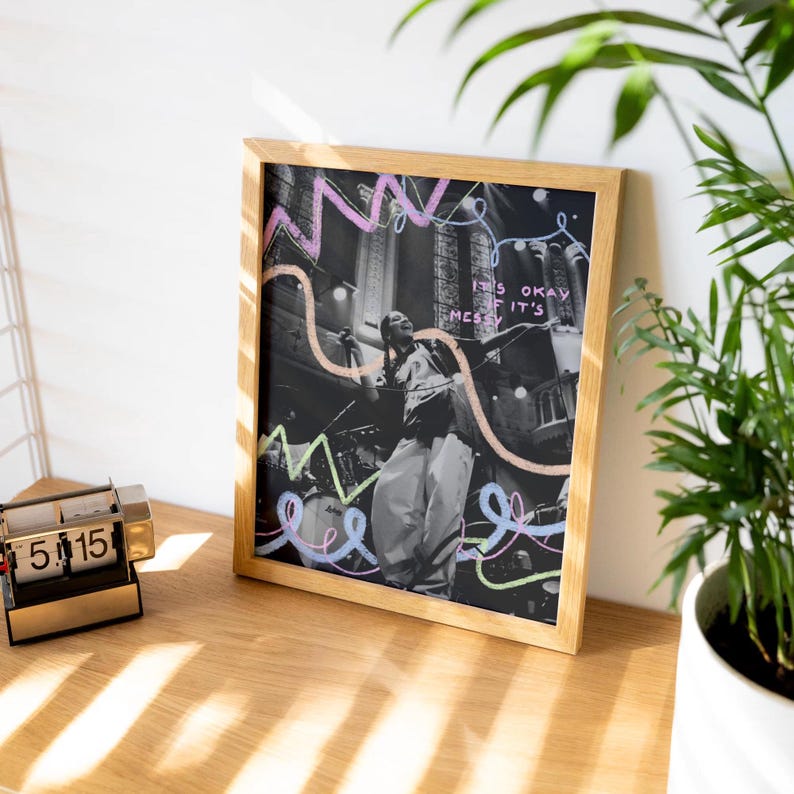 May include: A framed print featuring a black and white photograph of a person singing on stage. The background is a dark, blurred image of a crowd. The print has colourful lines and the text "It's okay if it's messy" written on it.