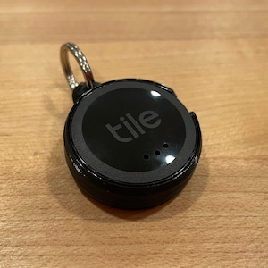 May include: A black Tile Bluetooth tracker with a silver key ring. The tracker has the word "tile" printed on it in white.