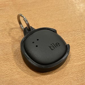May include: A black Tile Bluetooth tracker with a silver keyring. The tracker has a small hole with three dots and the word "tile" printed on it.