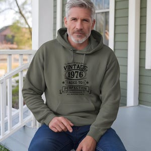 May include: An olive green hoodie with a front pocket and a drawstring hood. The hoodie features the text "VINTAGE 1976 AGED TO PERFECTION" in black lettering. The hoodie is a limited edition.
