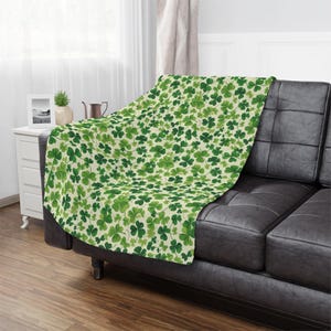 May include: A green and white shamrock-patterned throw blanket draped over a black leather couch. The blanket features a repeating pattern of green shamrocks on a cream-colored background. The blanket is soft and cozy, perfect for snuggling up on the couch.
