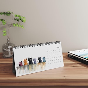 Peut inclure: Calendrier de bureau avec une illustration de sept chats colorés assis sur une clôture en bois. Le calendrier est ouvert sur le mois de mai, avec les jours du mois visibles. Le calendrier est posé sur une surface en bois.