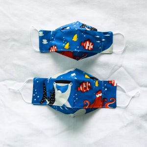May include: Two blue fabric face masks with a white background. The masks feature a colorful ocean-themed print with fish, an octopus, and a shark. The masks are made of cotton and have elastic ear loops.