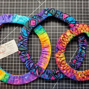 May include: Three colorful fabric steering wheel covers. The largest cover is rainbow with purple, blue, green, yellow, orange, and red. The middle cover is blue, black, and pink with geometric shapes. The smallest cover is purple, pink, orange, and yellow with a swirling pattern. The covers are on a black and white grid.