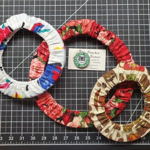 Pode incluir: Três pegadores de panela de tecido com padrões diferentes. O primeiro é branco com flores rosa, azuis e amarelas. O segundo é vermelho com flores rosa e brancas. O terceiro é bege com um padrão de xícara de chá marrom e verde. O cartão de visita diz "Stitchin' Sisters crafts & notions".