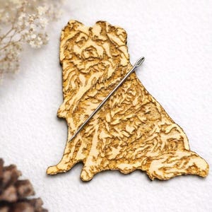 May include: A laser-cut wooden dog-shaped magnetic needle minder. The light brown wood has detailed etched patterns. A silver needle is attached. The text reads "Laser Cut Wooden Dog Magnetic Needle Minder" and "Custom Needle Minder."