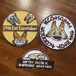 May include: Three embroidered patches featuring a cartoon Loth-cat from Star Wars. The first patch is yellow with the text "Loth Cat Caretaker" and a Loth-cat with its arms outstretched. The second patch is white with a red border and the text "Wookiee Love Loth-cat" and a Loth-cat with its mouth open. The third patch is brown with a white border and the text "Wookiee Love Loth-cat" and a Loth-cat lying down.