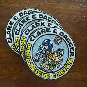 May include: A stack of round embroidered patches featuring Star Wars droids. The patches have a light blue background with the text "CLARK & DAGGER DROID RESCUE & REPAIR" in yellow and white. The patches depict R2-D2, BB-8, and other droids.
