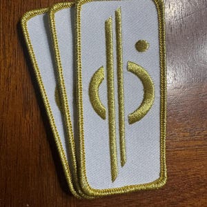 May include: Three white fabric patches with gold embroidered designs. The design features a stylized symbol with a circle and three vertical lines.