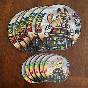 May include: A set of 7 round embroidered patches featuring a cartoon cat riding a robot with the word "REBEL" in rainbow colors and the word "PRIDE" in rainbow colors.