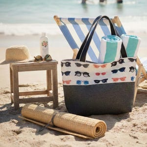 May include: A tote bag with a white upper section featuring a pattern of colorful sunglasses and a solid dark gray lower section. The bag contains two rolled teal and white striped towels and sits next to a straw hat, sunglasses, and a bottle.