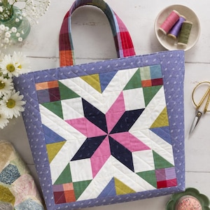 May include: A quilted tote bag featuring a central eight-pointed star pattern in shades of pink, navy blue, white, and green. The bag has a lavender border with a small white floral print and a multicoloured plaid handle. The stitching is visible.