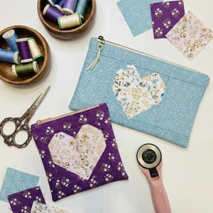 May include: A light blue quilted pouch with a floral heart design and a purple quilted pouch with a floral heart design. Wooden bowls hold spools of thread in various colors. A pink rotary cutter and scissors are also present.