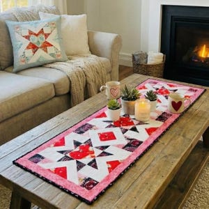May include: A rectangular quilted table runner featuring a geometric star pattern in shades of red, white, and black, with a pink floral border. The runner is decorated with three small potted succulents and two white ceramic mugs with heart motifs.