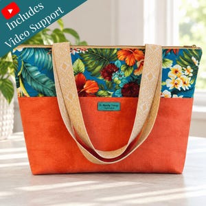 May include: A tote bag featuring a teal upper section with a vibrant tropical floral print and a solid orange lower section. It has two beige woven straps with a geometric pattern and a small teal label reading Be Mandy Things Handcrafted.