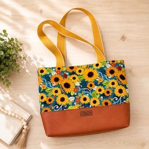 May include: A tote bag featuring a vibrant sunflower pattern on a teal background with yellow straps and a solid brown leather-like base. A small rectangular label with the text "Be Mandy Things" is centered on the brown bottom section.