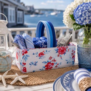 May include: A rectangular fabric caddy featuring a white background with a red and blue floral pattern. It has a sturdy, arched handle made of blue-toned fabric strips and contains folded blue-patterned napkins and silver metal cutlery.