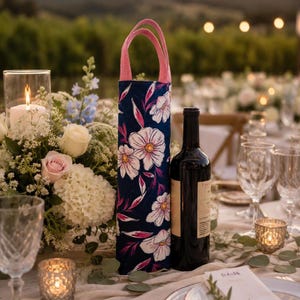 May include: A tall, cylindrical fabric wine bottle gift bag featuring a dark navy blue background with a white and pink floral pattern and two pink fabric handles, positioned next to a dark glass wine bottle on a table setting.