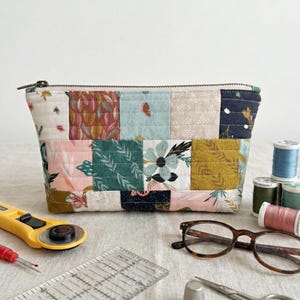 May include: A patchwork fabric cosmetic bag with a zipper closure. The bag features a variety of floral and geometric patterns in shades of pink, green, and gold. Sewing tools, glasses, and spools of thread are arranged around the bag.