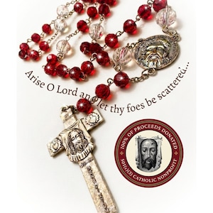 May include: A rosary featuring alternating translucent red and clear faceted beads connected by metal links. It includes a silver-toned crucifix with a central face relief and the text Arise O Lord and let thy foes be scattered.