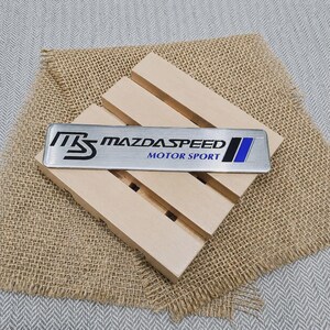 Puede incluir: Un emblema metálico rectangular de color plateado que presenta el logotipo MS estilizado en negro, el texto MAZDASPEED MOTOR SPORT en negro y azul, y una franja vertical negra y azul en el lado derecho, colocado sobre una paleta de madera clara.