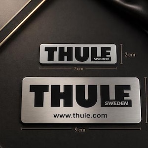 May include: Two rectangular silver metal plates with black text. The smaller plate measures 7 inches by 2 inches and reads THULE SWEDEN. The larger plate measures 9 inches by 4.5 inches and reads THULE SWEDEN with the website www.thule.com below.