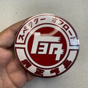 May include: A circular emblem featuring a deep red center with a large white stylized character. The outer white border contains red Japanese text, including the katakana characters for "specter" and "offroad" along with other Japanese script.