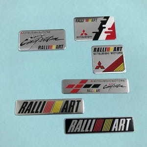 May include: A collection of silver and black Ralliart car emblems. The emblems feature the Ralliart logo in various designs, with red, yellow, and black stripes. Some emblems include the Mitsubishi logo and text.