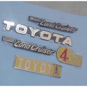 Op de afbeelding: Een verzameling van vijf auto-emblemen, waaronder twee zilveren Toyota Land Cruiser-badges, een groot zilveren Toyota-logo, een rechthoekig geel Toyota-embleem en een ovaal embleem met een rood cijfer 4 en de tekst 4 WHEEL DRIVE.