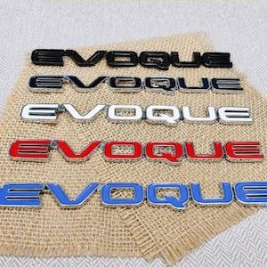 May include: Five individual car emblems featuring the word EVOQUE in capital letters. The emblems are arranged in a vertical stack, each displaying a different color finish: solid black, black with silver trim, white with silver trim, red with silver trim, and blue with silver trim.