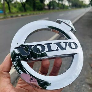 May include: A chrome Volvo emblem with the brand name in black lettering. The emblem is circular with a rectangular top piece. The background is a blurred view of a road and trees.