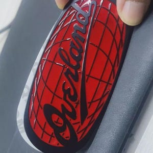 May include: Oval-shaped emblem with a red background featuring a black grid pattern and the word "Overland" in a cursive font. The emblem has a black border.