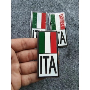 Op de afbeelding: Rechthoekige emblemen met de kleuren van de Italiaanse vlag: groen, wit en rood. Het woord "ITA" staat in witte letters onderaan. Drie emblemen zijn zichtbaar, waarvan er één in een hand wordt gehouden.