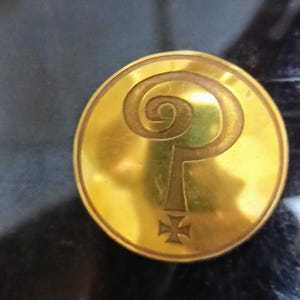May include: A gold-coloured circular coin featuring a raised question mark design and a cross at the base. The coin has a shiny surface and is set against a dark background.
